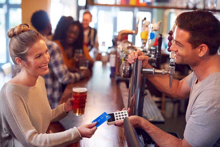 Smiling Female Customer In Bar Making Contactless Payment With Card For Drinks To Male Bartender
